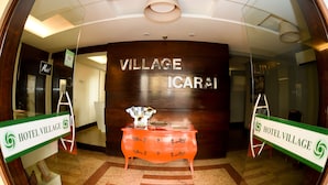 Un área de recepción con una pared de madera, un letrero que dice "VILLAGE Icarai" y un aparador vintage naranja con un florero y revistas.