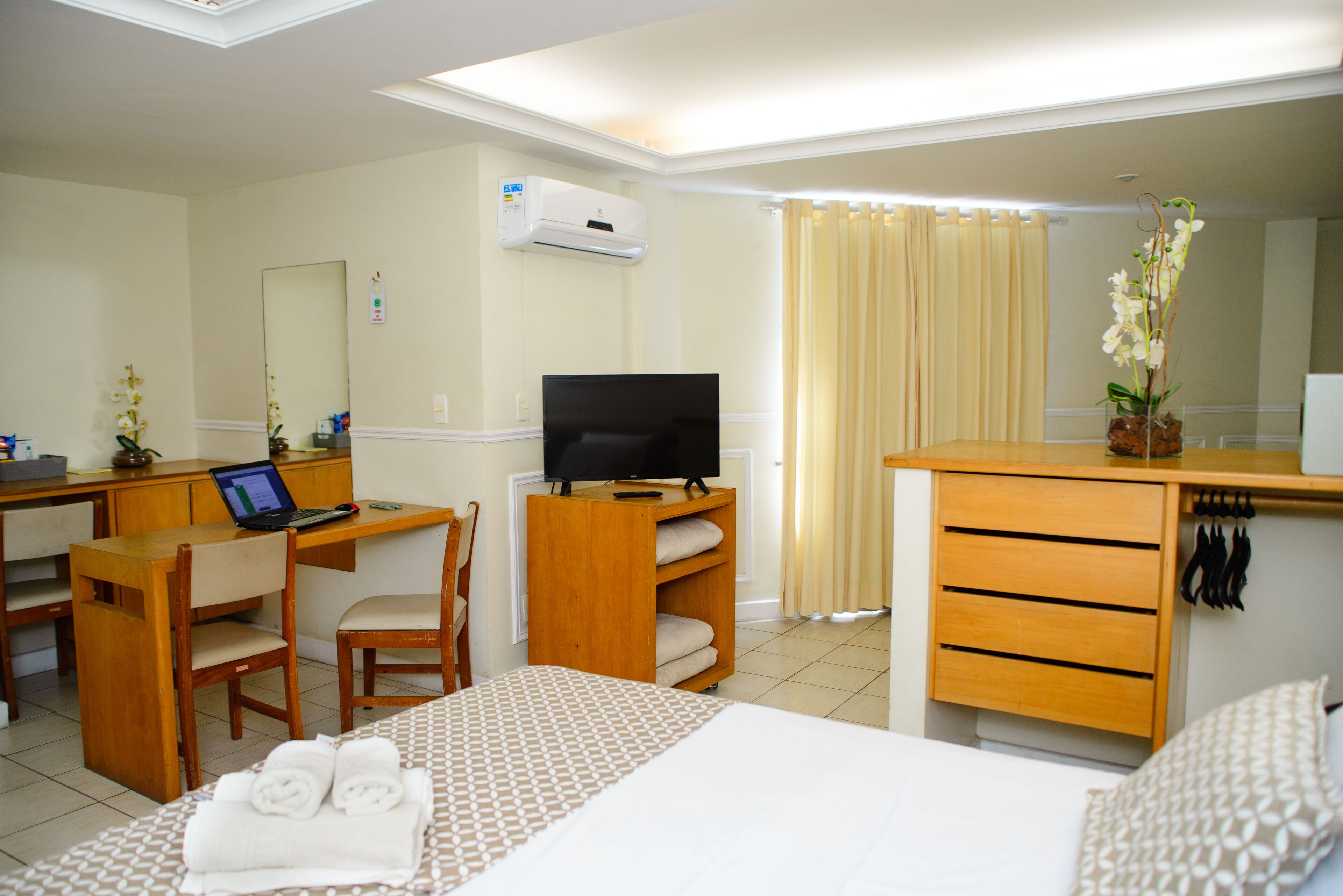 Habitación de hotel con cama, escritorio con computadora portátil, televisor, espejo, cómoda y zona de ducha.