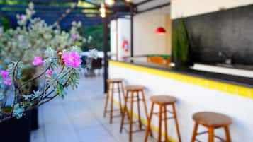 Uma área de bar com bancos de madeira e balcão, rodeada por plantas em vasos com flores cor-de-rosa.