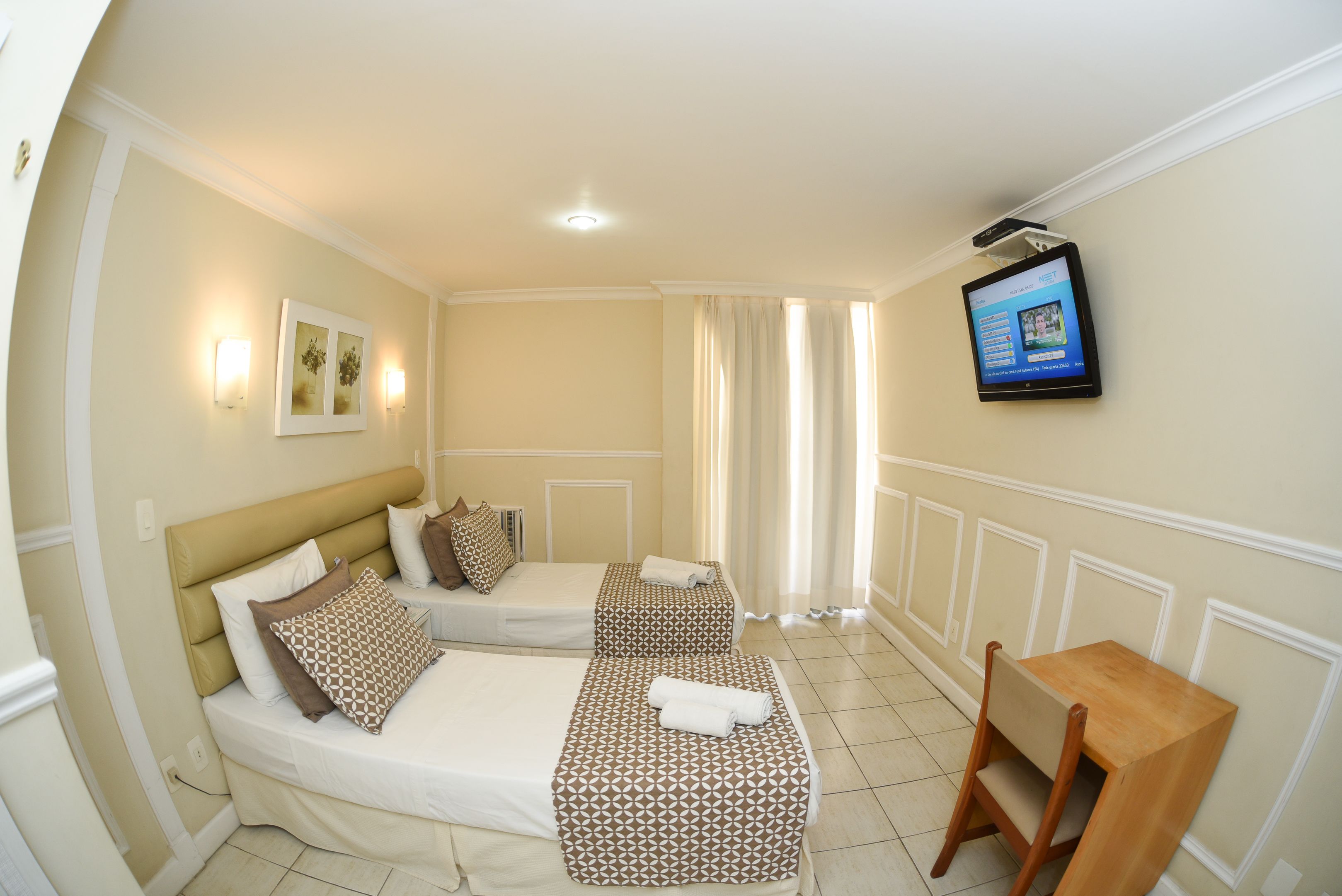Quarto de hotel com duas camas, uma TV fixada na parede e uma pequena mesa de madeira.
