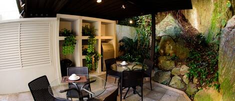 Una mesa con superficie de vidrio y cuatro sillas, rodeada de plantas en macetas y un muro de piedra.