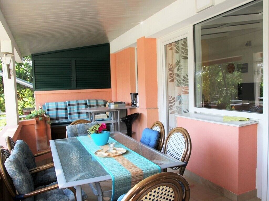 A dining area with a table set for two, surrounded by wicker chairs. A sofa and a small table with a coffee machine are visible in the background.