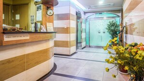 Zona de recepción de hotel con un mostrador de madera, un reloj y un letrero que indica un restaurante.