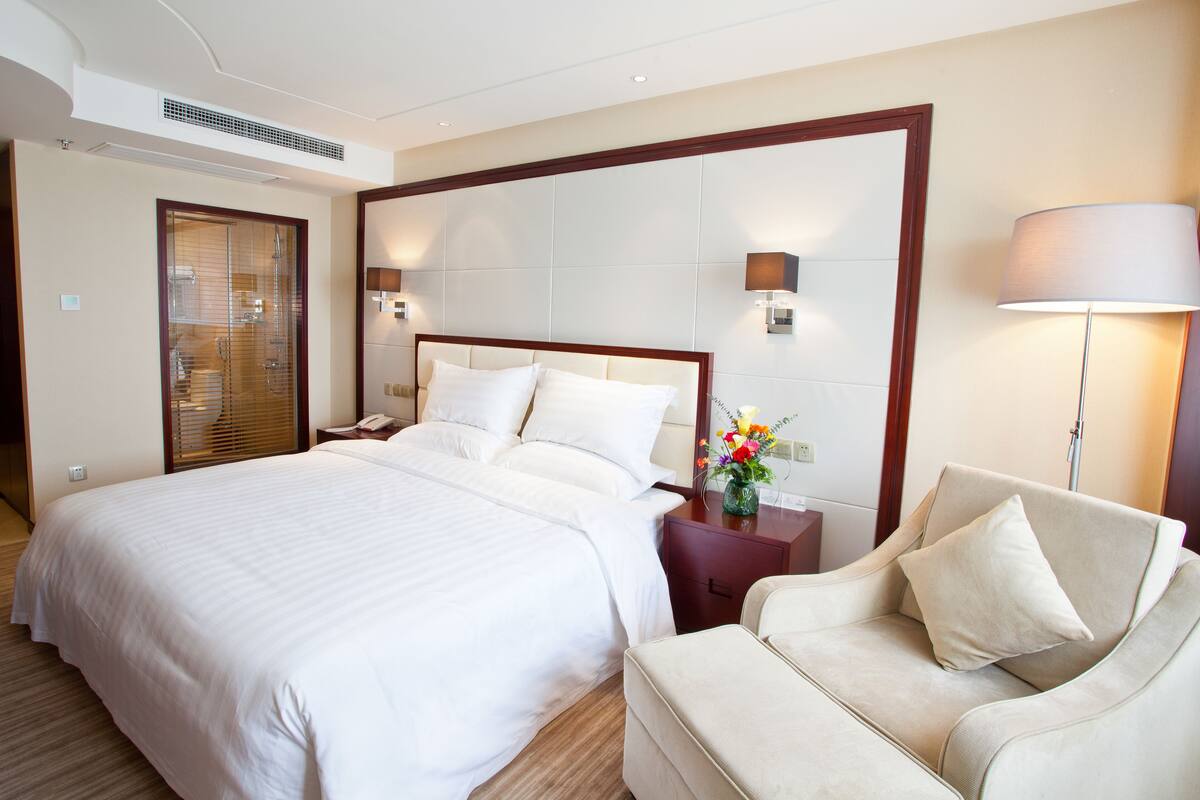 A hotel room with a large bed, a bedside table with a lamp, a sofa, and a shower area.