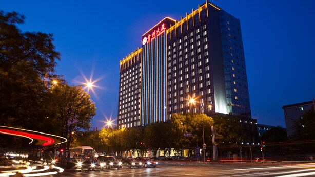 Um hotel alto com uma placa grande no topo, vários andares e um cruzamento movimentado em primeiro plano, com rastros de luz de veículos em movimento.