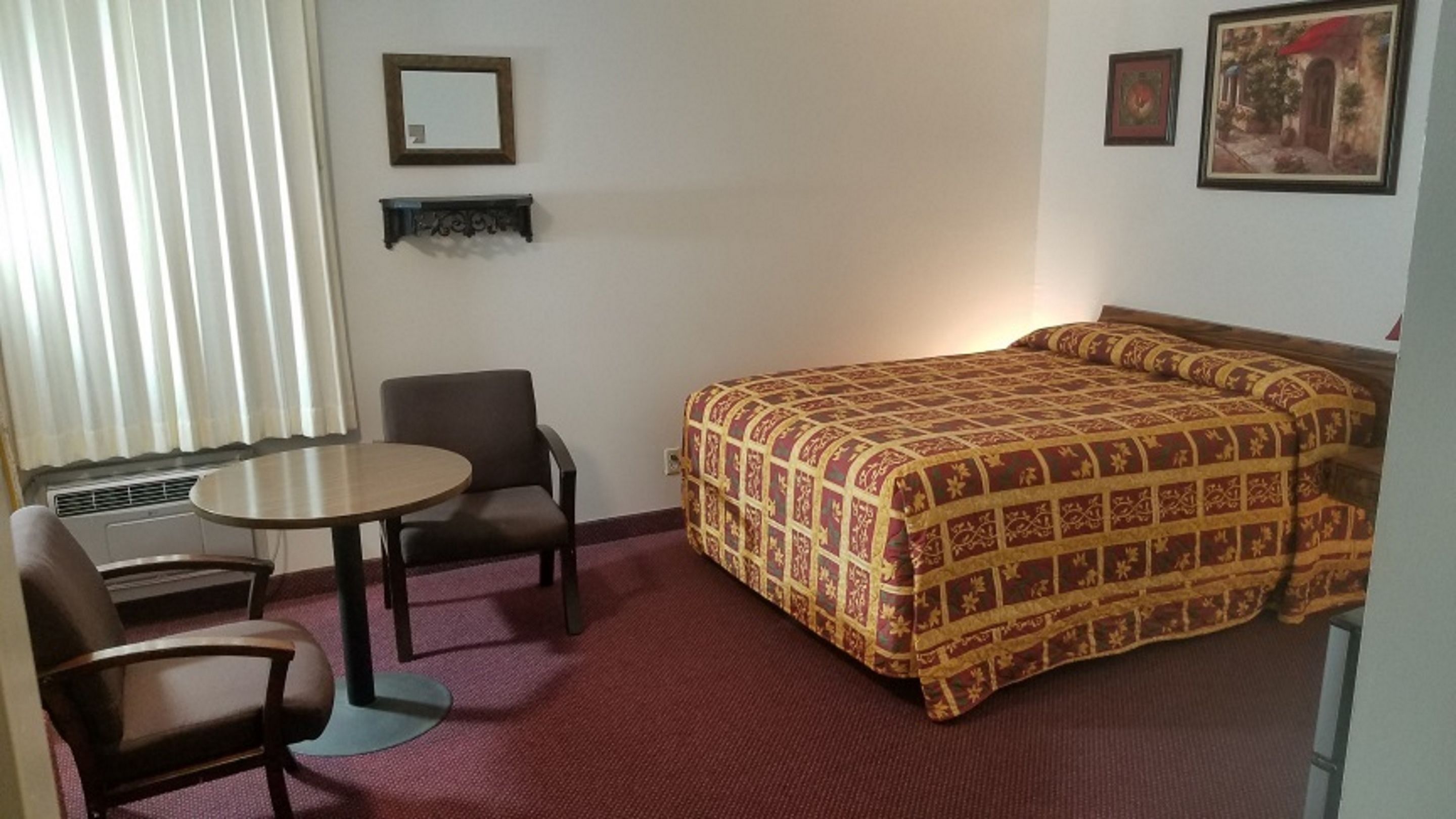 A hotel room with a bed, a small table, two chairs, a mirror, and two framed pictures on the wall.