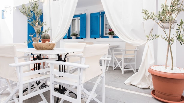 A white outdoor seating area with tables and chairs, potted plants, and blue shutters on the windows. - Villa Iokasti (Santorini)