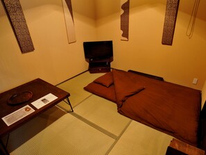 A traditional Japanese-style room with tatami flooring, a low wooden desk, a television, and a bed with brown bedding. - Theatre and Library Residence -Kyoto Murasakino- (Kyoto)