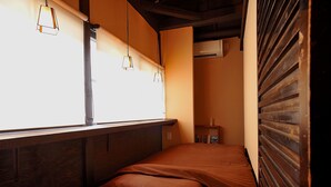 A small, cozy room with a bed, pillows, a shelf with books, and a window with hanging lights.