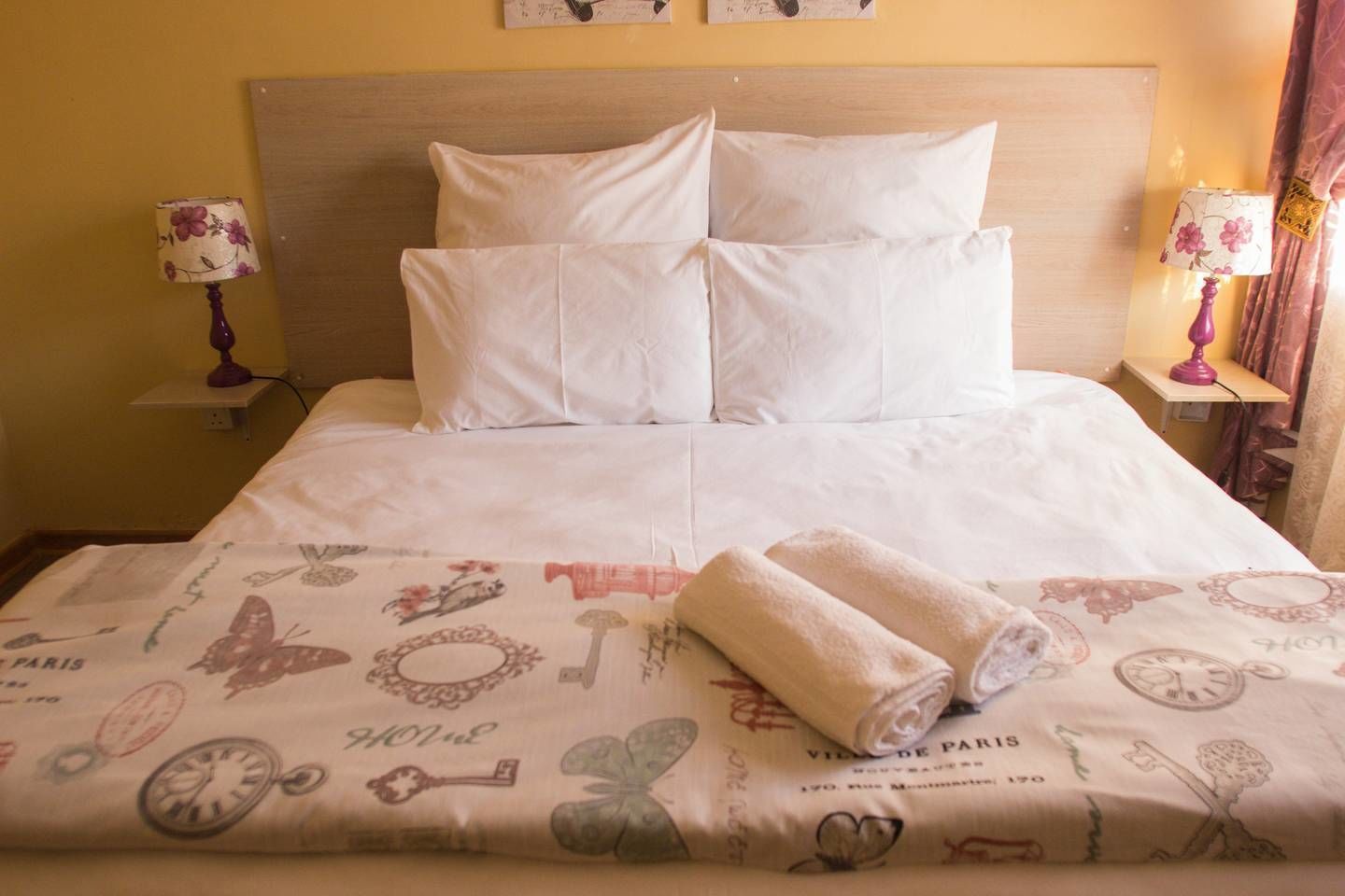 A neatly made bed with white linens and a Paris-themed duvet cover, two bedside lamps, and a wooden headboard.