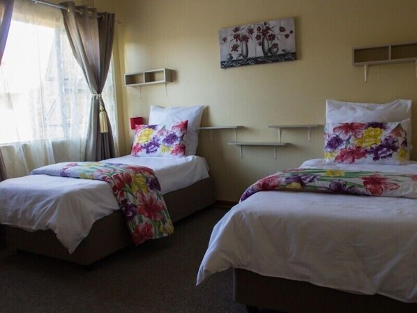 A hotel room with two beds, each with floral bedding and white pillows. There is a painting on the wall above the beds, and a small shelf to the right.