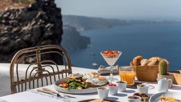 Una mesa con alimentos para el desayuno, incluyendo tostadas, fruta y un vaso de zumo de naranja, con vistas a un paisaje costero.