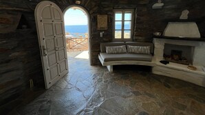 A stone-floored room with a stone fireplace, a sofa, and a view through an open door to the sea. - The Stone Windmill (Kea)