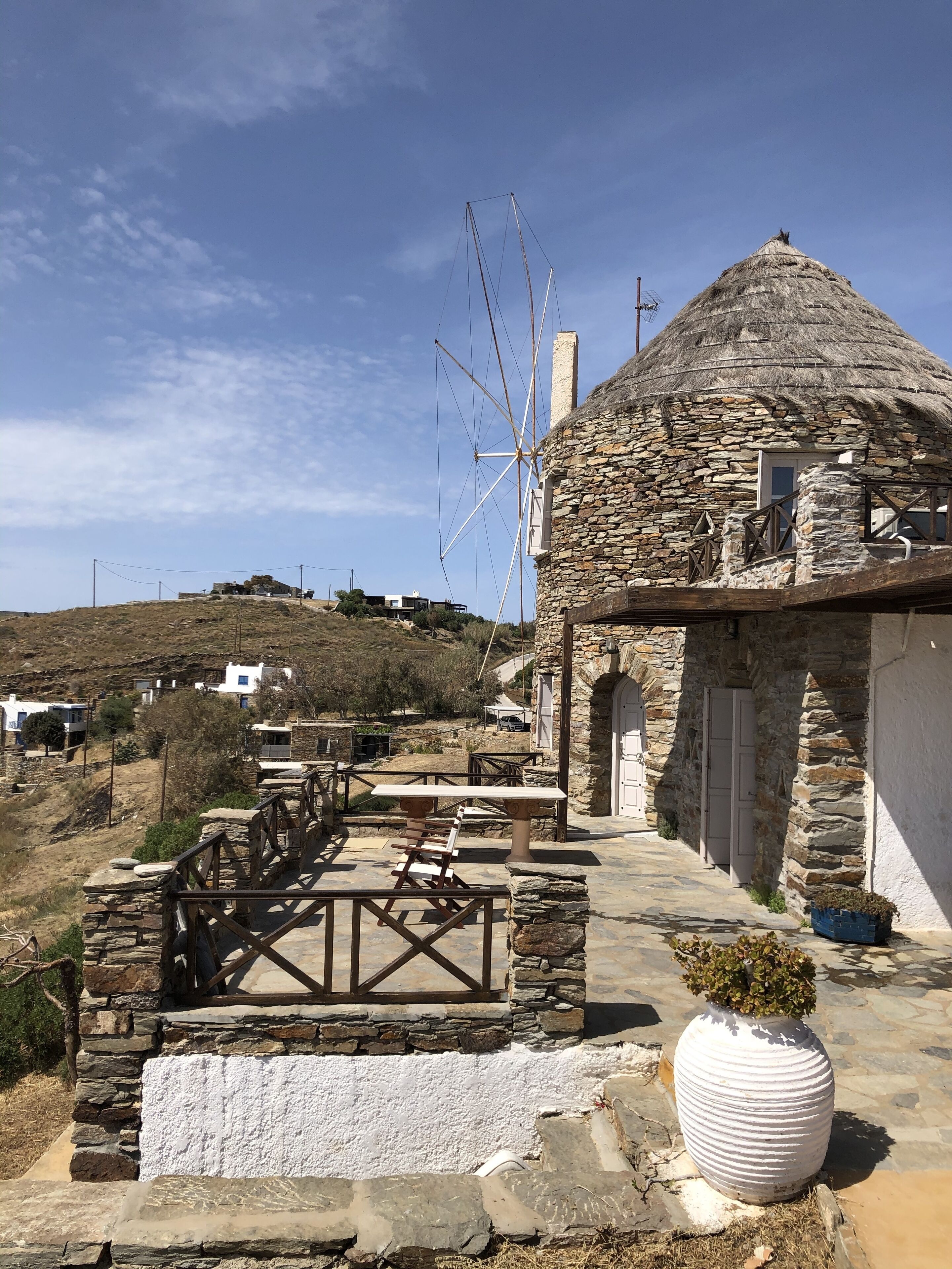 A stone building with a thatched roof, a windmill, and a paved patio with tables and chairs.