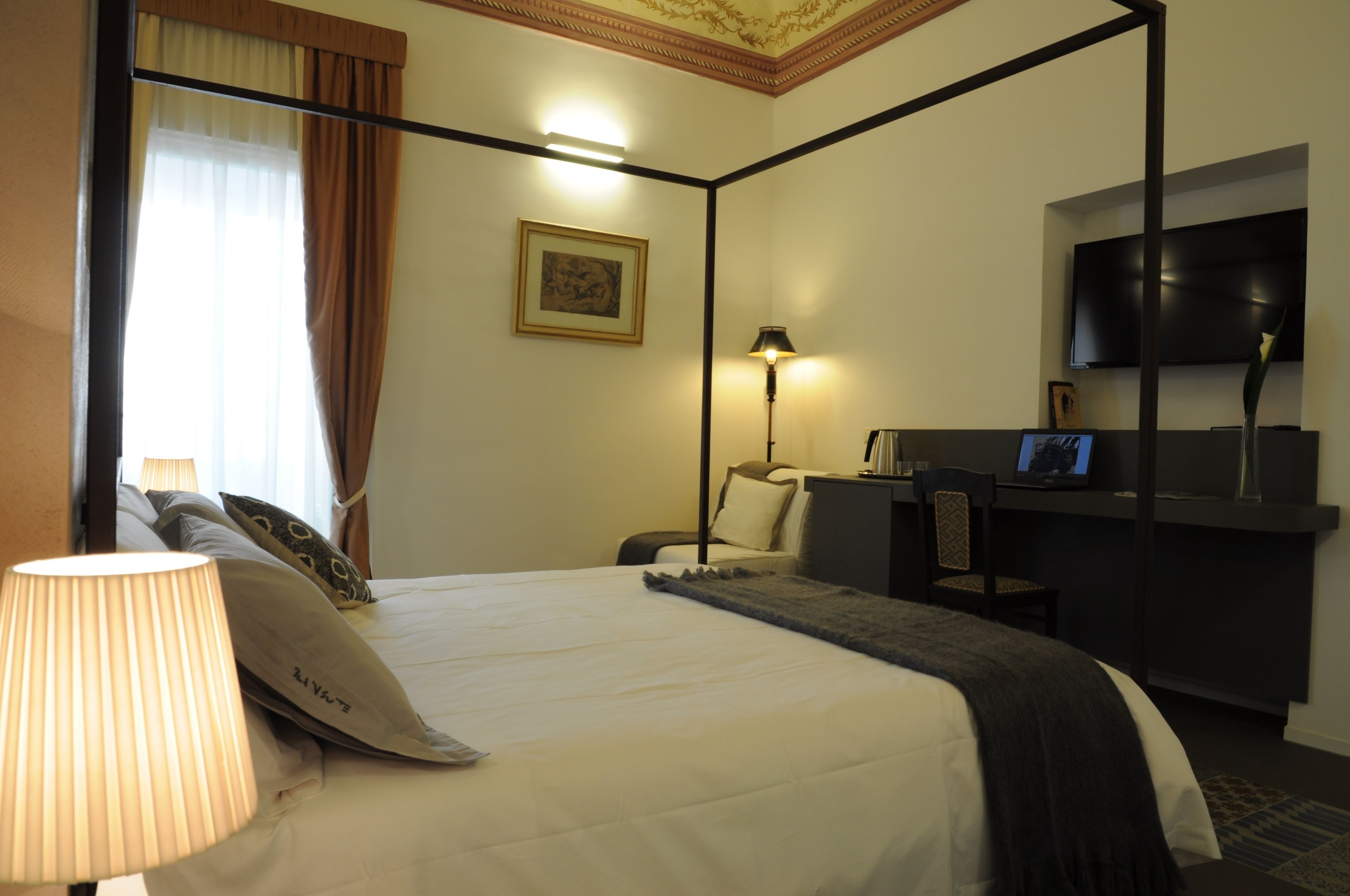 Habitación de hotel con cama con dosel, un escritorio con silla, un televisor de pantalla plana montado en la pared y una lámpara en la mesita de noche.