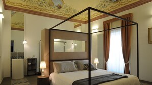 A four-poster bed with a mirror above it, a bedside table with a lamp, and a view of the bathroom through an open door. - Iblainsuite (Ragusa)