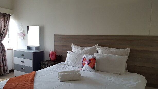 A neatly made bed with white bedding and a single pillow, a wooden headboard, a bedside table with a red vase, and a dresser with drawers. - Lolos Elegant Three Bedroom Apartment (Harare)