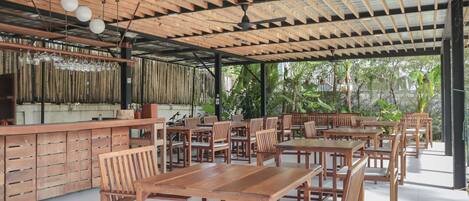 Un área de comedor al aire libre techada, con mesas y sillas de madera, una barra con vasos y vistas a zonas verdes.