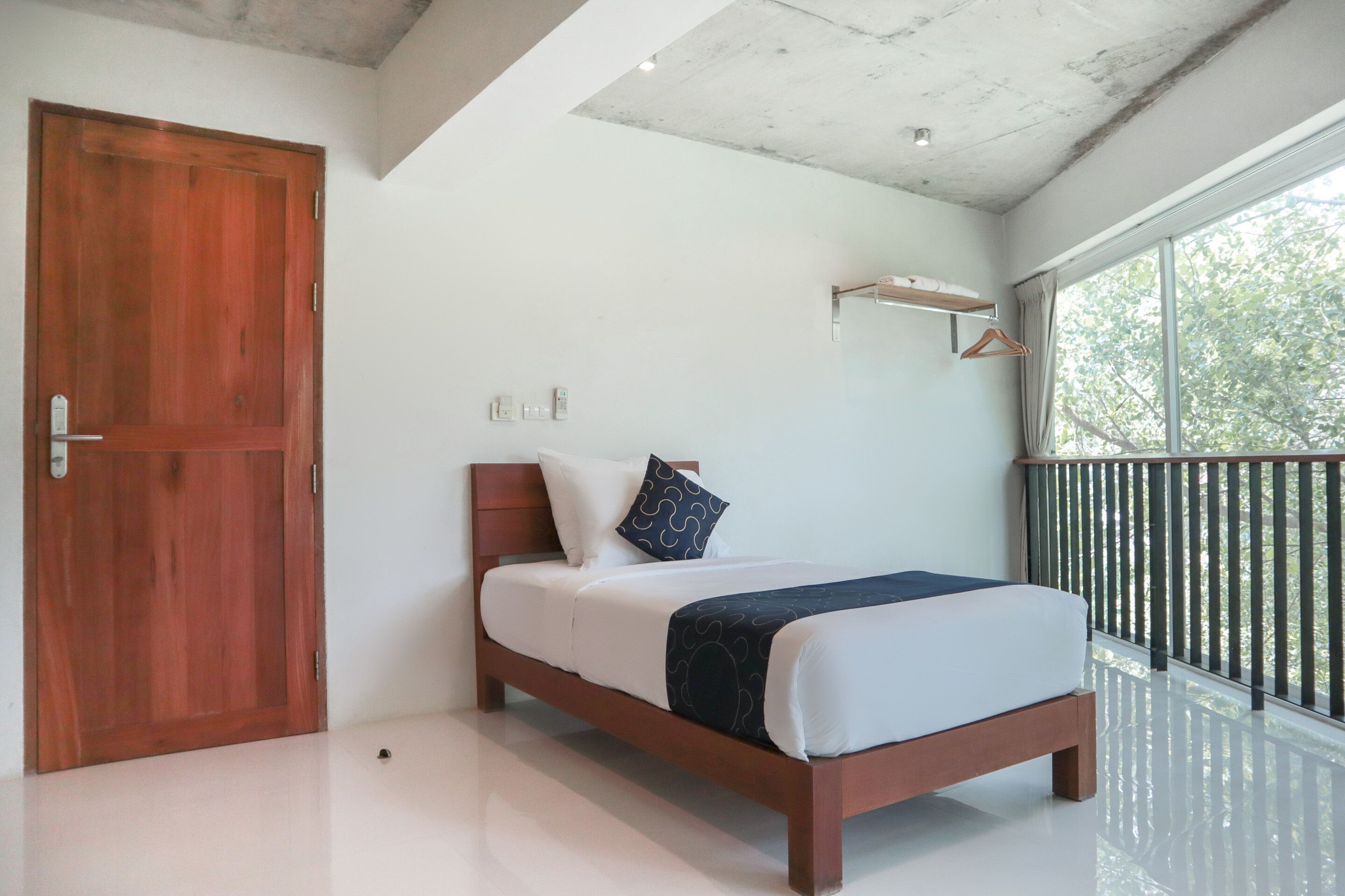 A bedroom with a wooden bed, white bedding, a black and white pillow, a small shelf, and a large window with a view of trees.