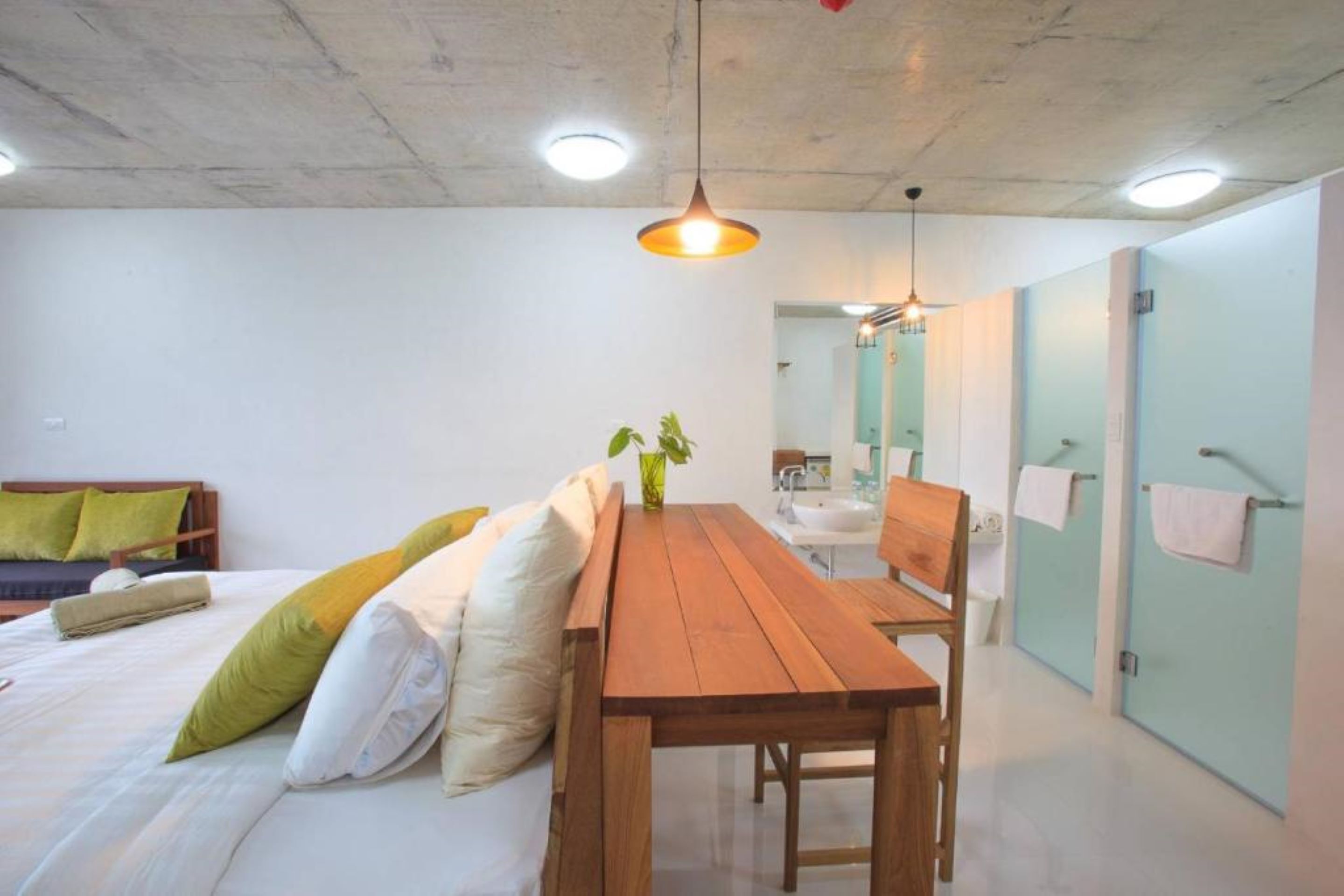 A modern bedroom with a wooden bed, a wooden table, and a bench. There is a small plant on the table. The room has a concrete ceiling and light fixtures.