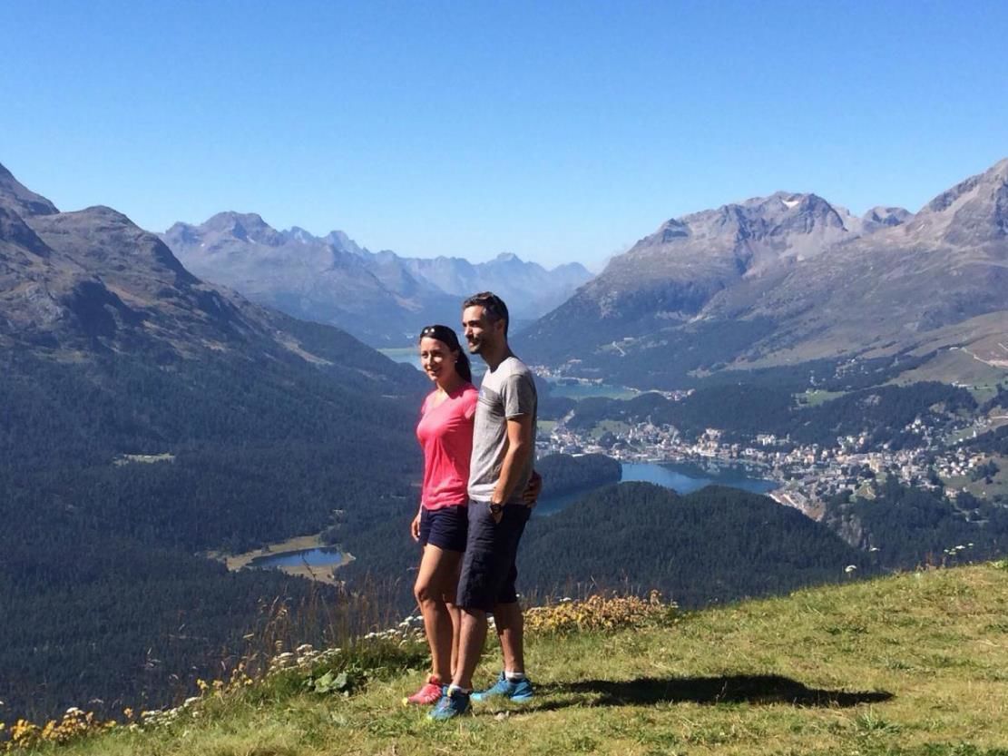 A couple standing on a grassy hill with a mountainous landscape in the background.
