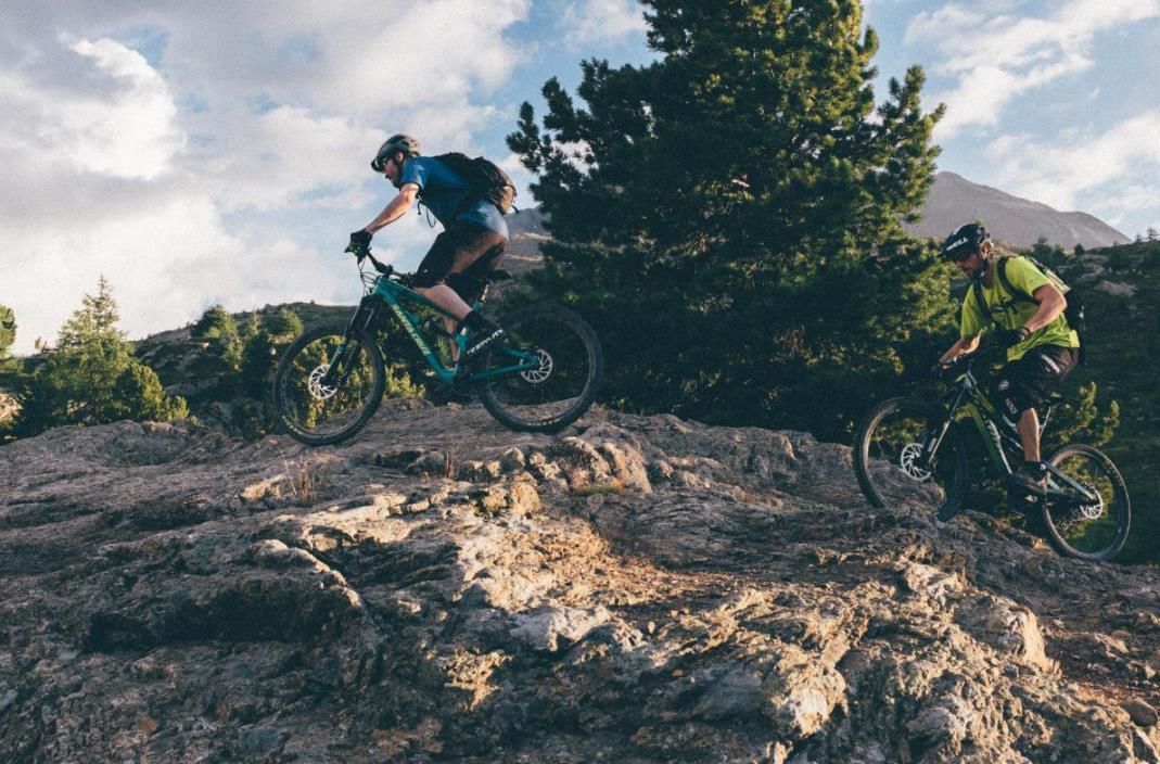 Two mountain bikers on a rocky trail with trees and a mountain in the background.