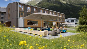 A modern hotel with a covered outdoor seating area, surrounded by a garden with yellow flowers and a mountainous backdrop. - Bever Lodge (Bever)