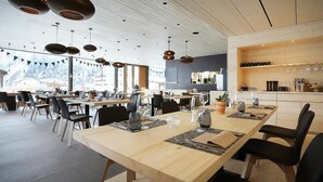 A modern dining area with wooden tables and black chairs, large windows with a view of mountains, and pendant lighting. - Bever Lodge (Bever)