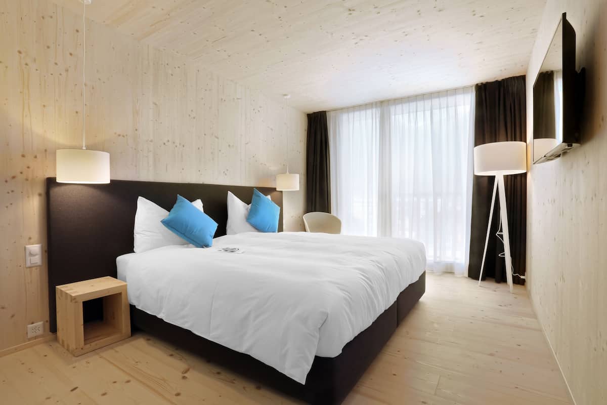 Quarto de hotel moderno com uma cama grande, abajures, uma pequena mesa de cabeceira de madeira e uma janela ampla com cortinas.