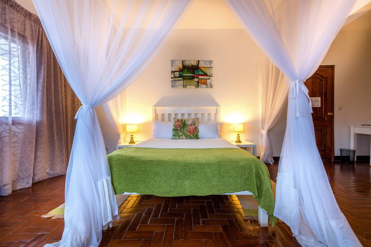 A bedroom with a bed, green bedspread, white curtains, and a painting on the wall.