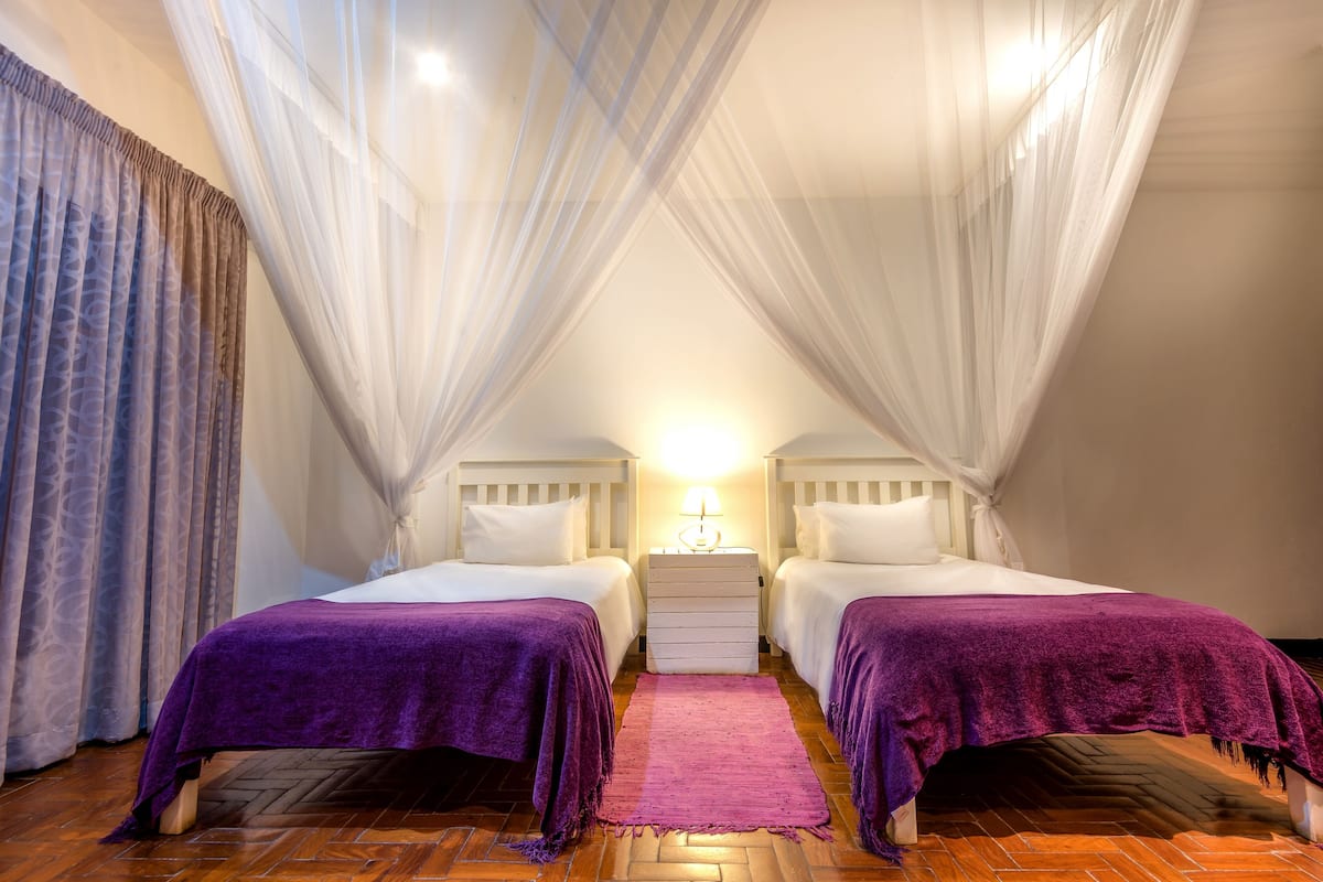 A bedroom with two beds, a wooden floor, a purple rug, and sheer curtains.