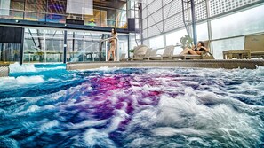 A spa pool with a waterfall feature, lounge chairs, and a glass-enclosed area with a view of the city.