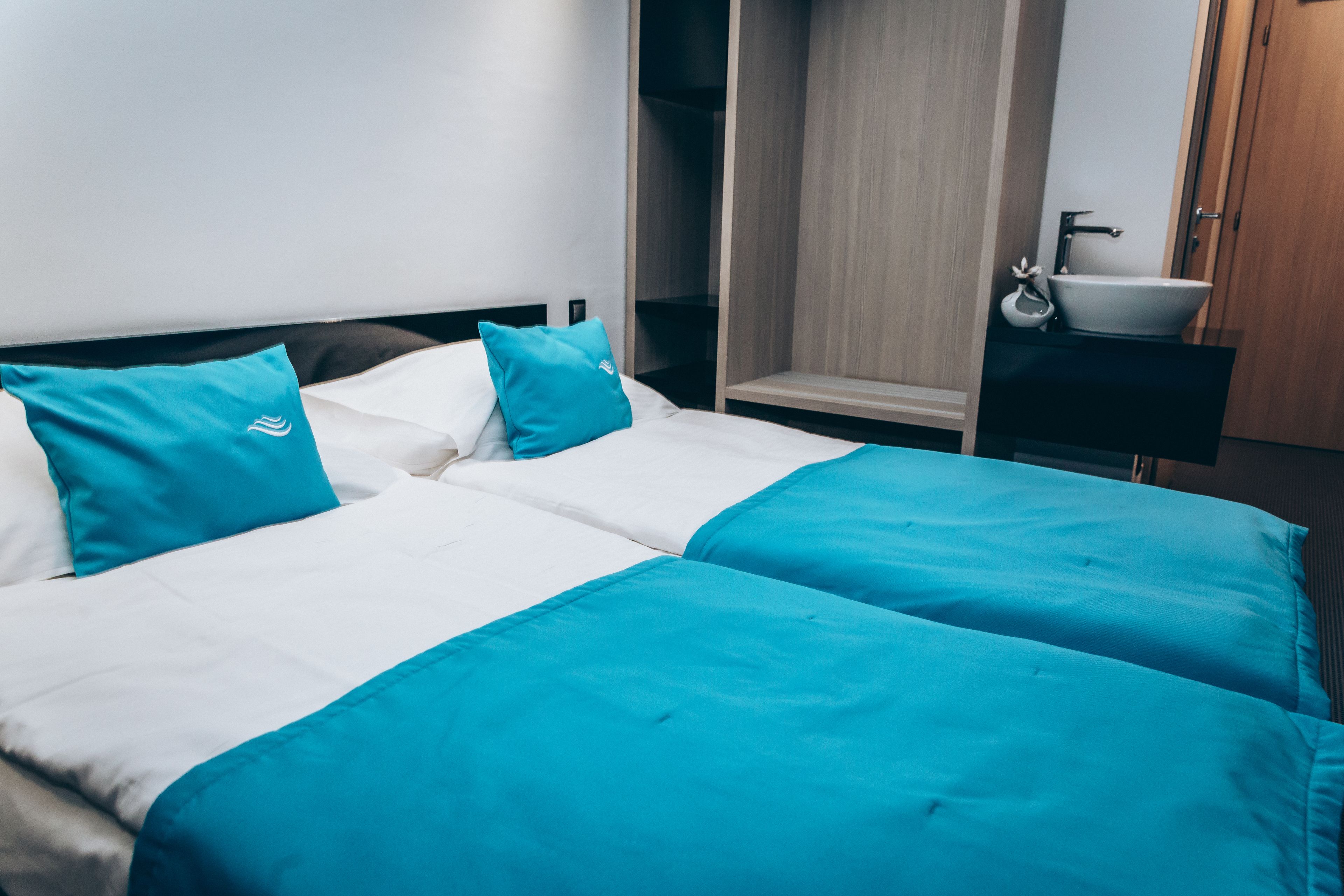 A hotel room with two beds, each with white and teal bedding, a wooden wardrobe, and a small bathroom with a sink and mirror.