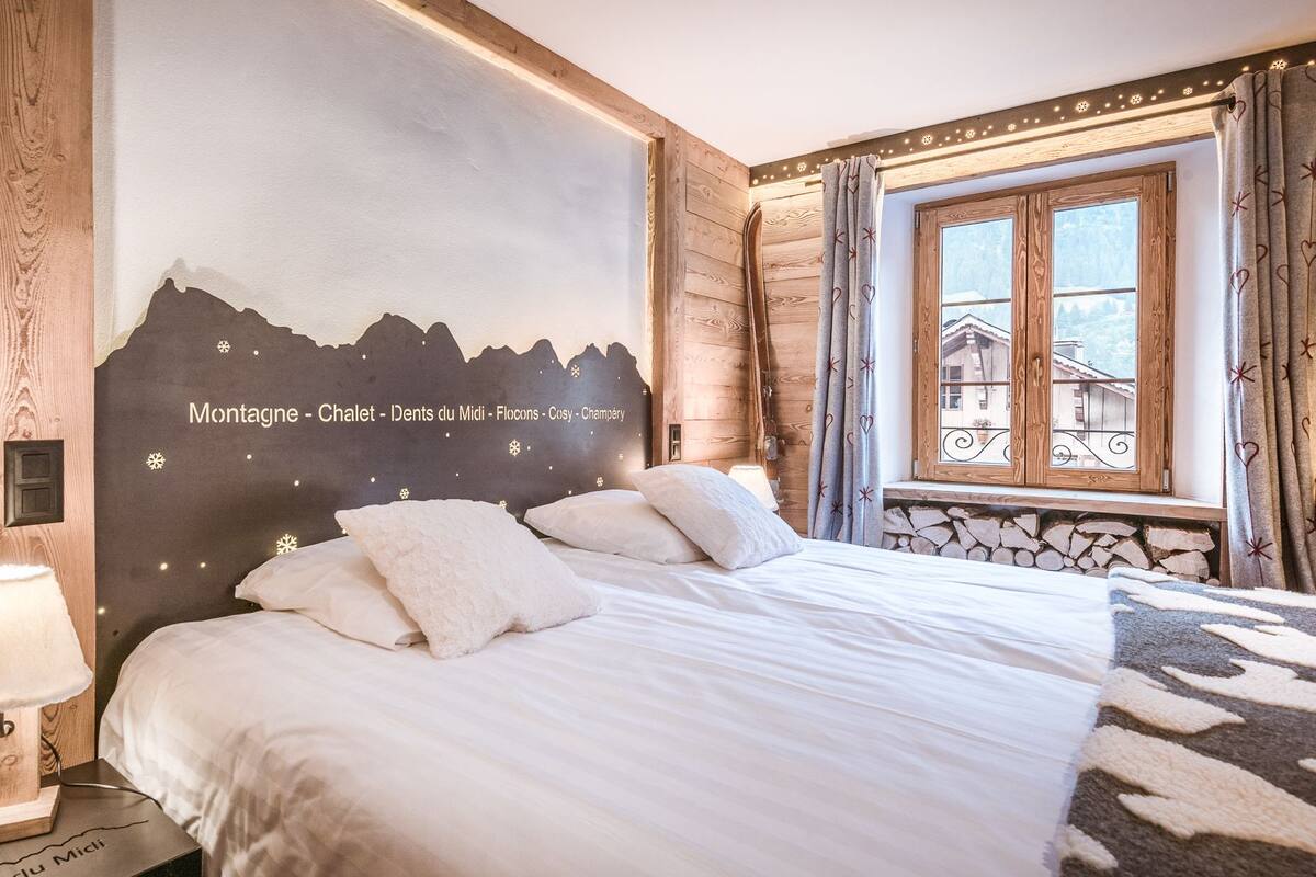 A cozy hotel room with a bed, a nightstand, a lamp, a window with curtains, and a wall mural depicting a mountain scene.