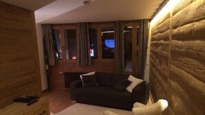 Una habitación acogedora con detalles de madera, un sofá negro, una silla blanca y una ventana con cortinas.