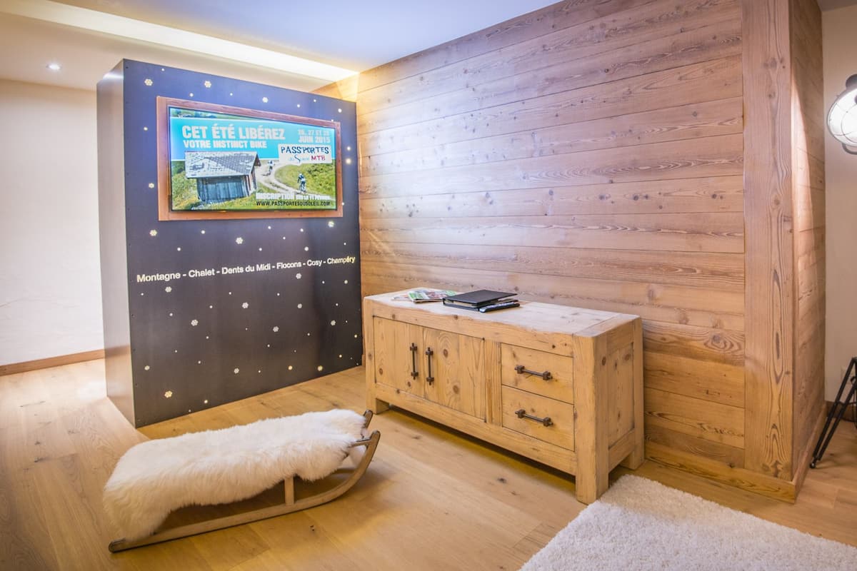 A room with a wooden floor, a white fur bench, a wooden cabinet, a desk with a laptop, a standing lamp, and a wall with a poster.