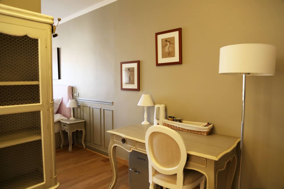 Ein Hotelzimmer mit einem Holztisch, einem Stuhl, einer Lampe, einem kleinen Tisch ebenfalls mit Lampe, einem Bett mit einem rosafarbenen Kopfkissen und zwei gerahmten Bildern an der Wand.