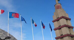 A row of flags on flagpoles, a building with a tower, and a clear blue sky. - Les Suites de Vanves (Vanves)