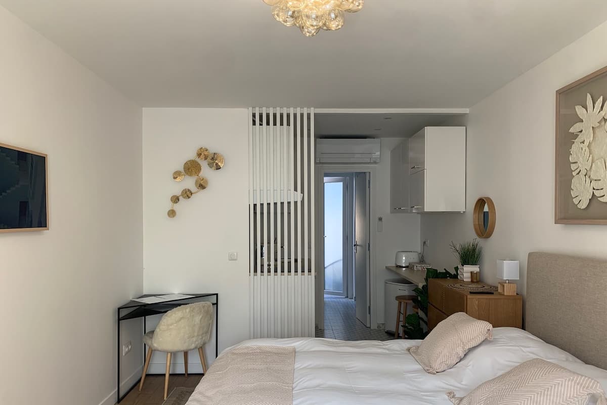 Ein modernes Schlafzimmer mit einem großen Bett, einem Schreibtisch und einem Sessel. Eine Tür führt zu einem Badezimmer und einer Küchenzeile.