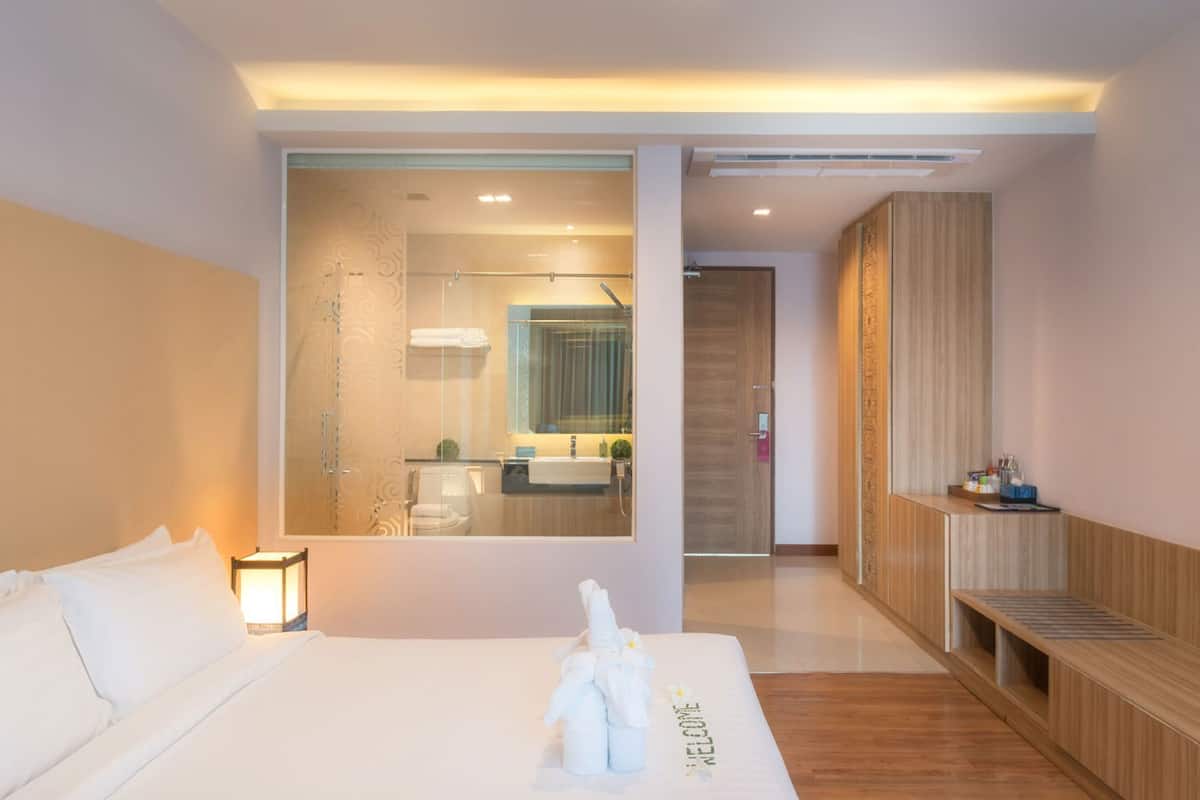 Hotellihuone, jossa on suuri sänky, yöpöytä lampun kanssa, peili, puinen vaatekaappi ja kylpyhuone, joka on näkyvissä lasiseinän läpi.