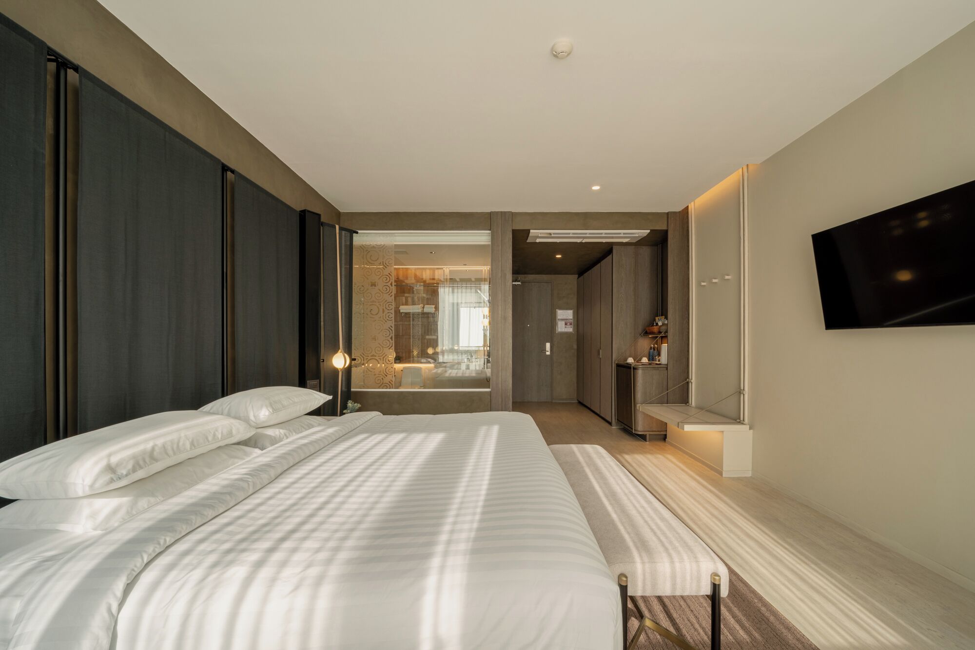 A modern hotel room with a large bed, a TV mounted on the wall, and a bathroom visible through a glass door.