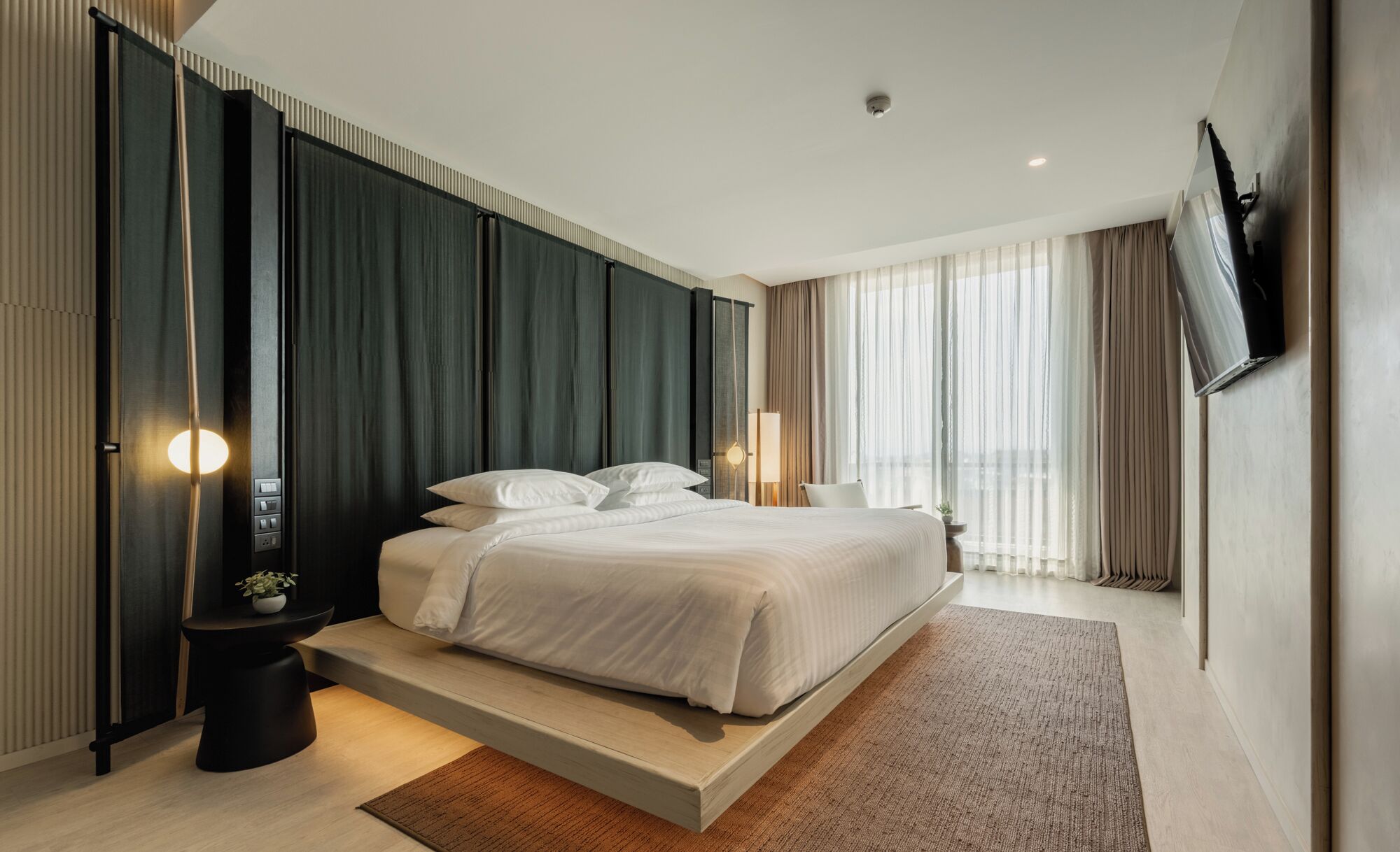 Une chambre d’hôtel moderne dotée d’un grand lit, d’une table de chevet, d’une commode, d’un téléviseur fixé au mur et d’une fenêtre avec des rideaux.