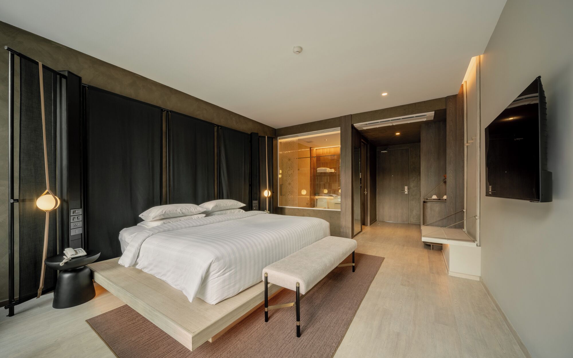 Een moderne slaapkamer met een groot bed, nachtkastjes, een bankje, een televisie en een badkamer zichtbaar door een geopende deur.