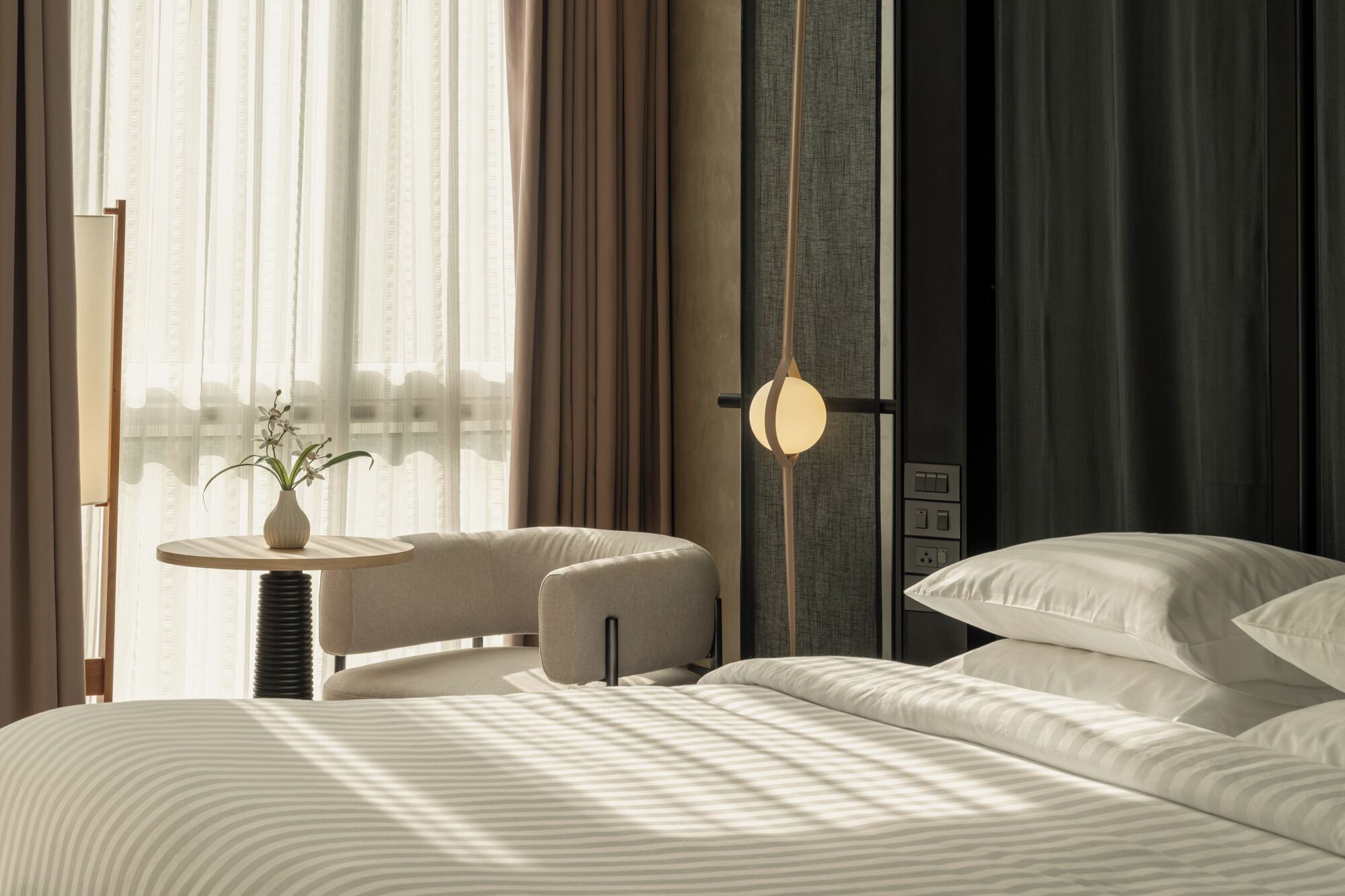 A modern hotel room with a bed, a small round table with a vase of flowers, a chair, and a unique wall-mounted light fixture.