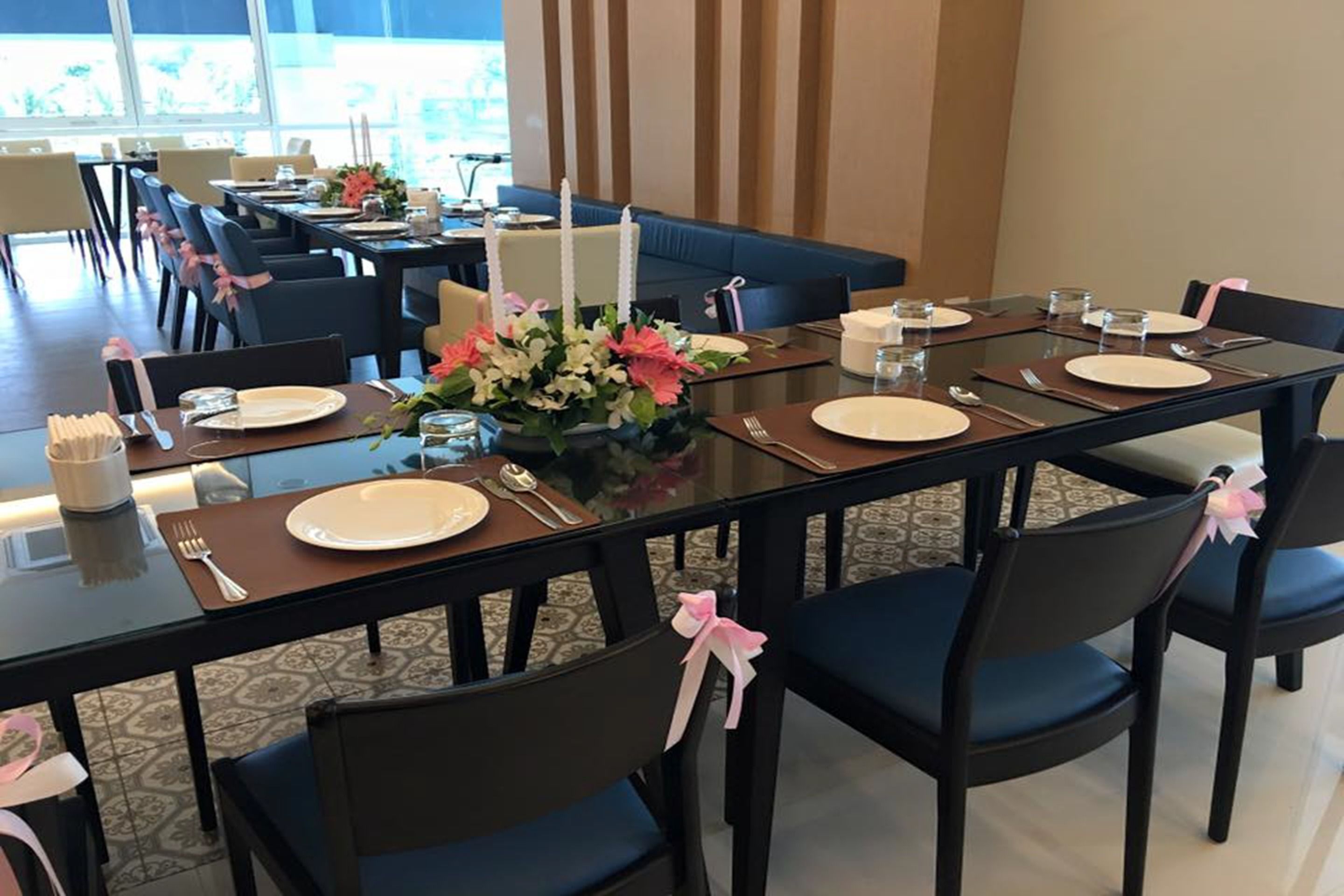 A dining area with tables set for four, each with a white plate, cutlery, and a centerpiece. Chairs are upholstered in dark fabric with pink bows.