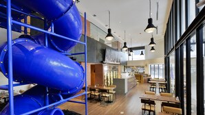 Un espacio interior moderno con una zona de juegos que incluye toboganes azules, mesas y sillas de madera dispuestas para comer o socializar.