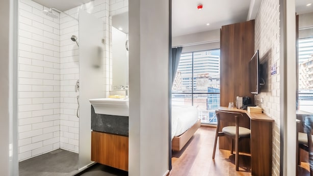 Habitación de hotel moderna con ducha, lavamanos, cama, escritorio y un amplio ventanal con vistas a la ciudad.