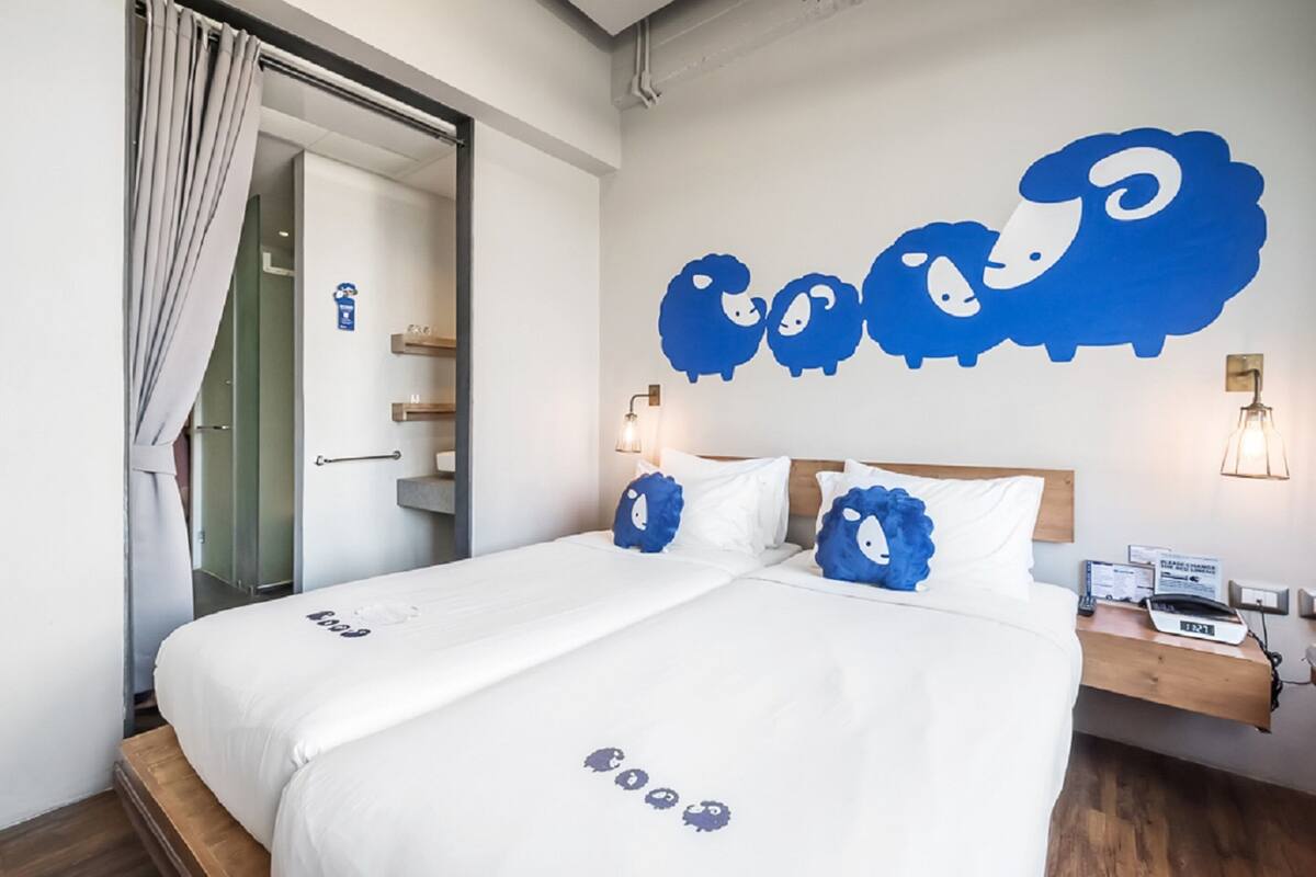A double bed with white bedding and blue decorative pillows, a wooden headboard, and a nightstand with a lamp and a device.