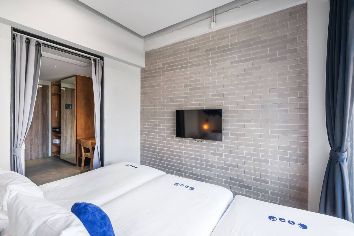 A hotel room with a brick wall, a flat-screen TV mounted above a bed, and a wooden desk with a chair.