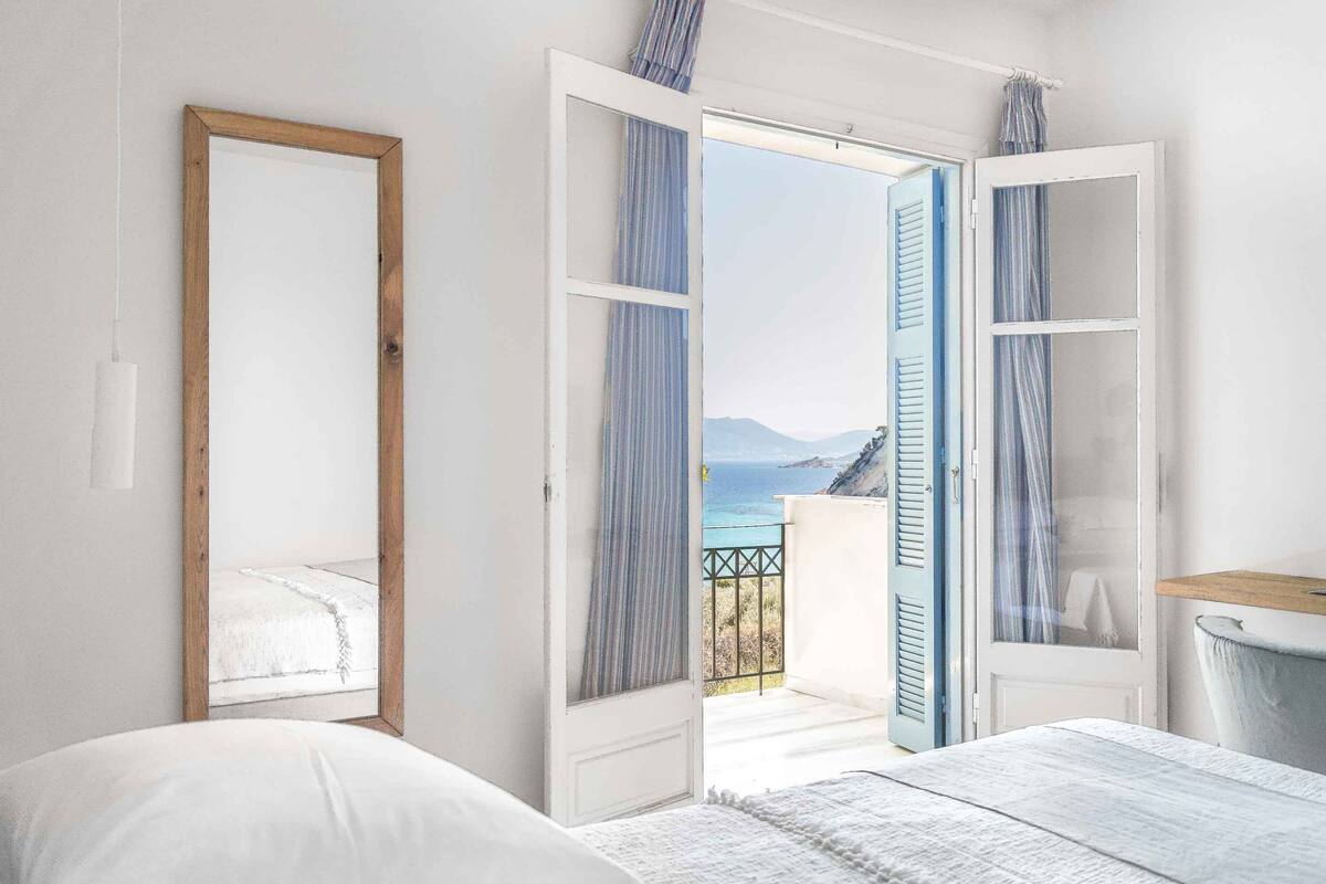 Un dormitorio con cama, un espejo grande y una puerta abierta que da a un balcón con vista al mar.